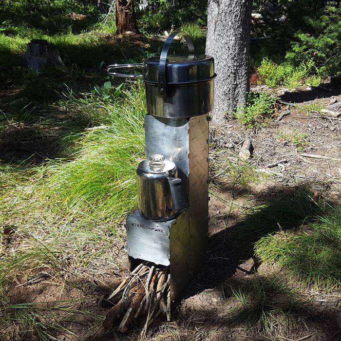A tall StealthFyre rocket stove tower with a pot on top and a percolator on a side shelf, in open pine forest